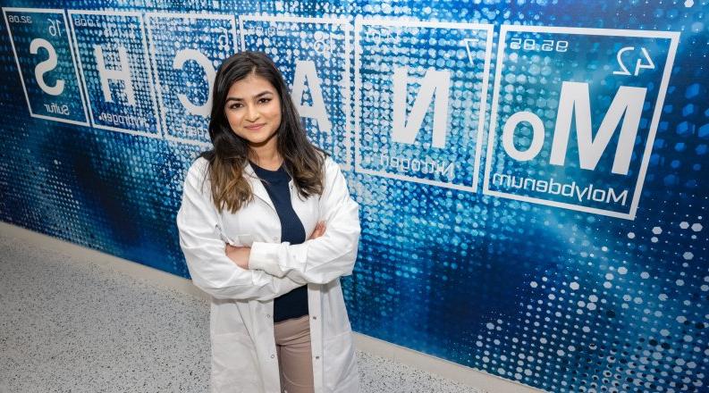 Antra Patel wears a lab coat and stands in front of a sign made up of symbols from the Periodic Table of Elements and spelling “MoNArCHS.”