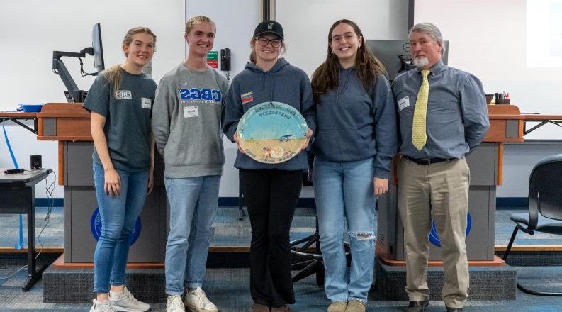 A group of students and their teacher stand with an oval-shaped bowl recognizing their first-place finish in the first-ever Ocean Bowl competition.  